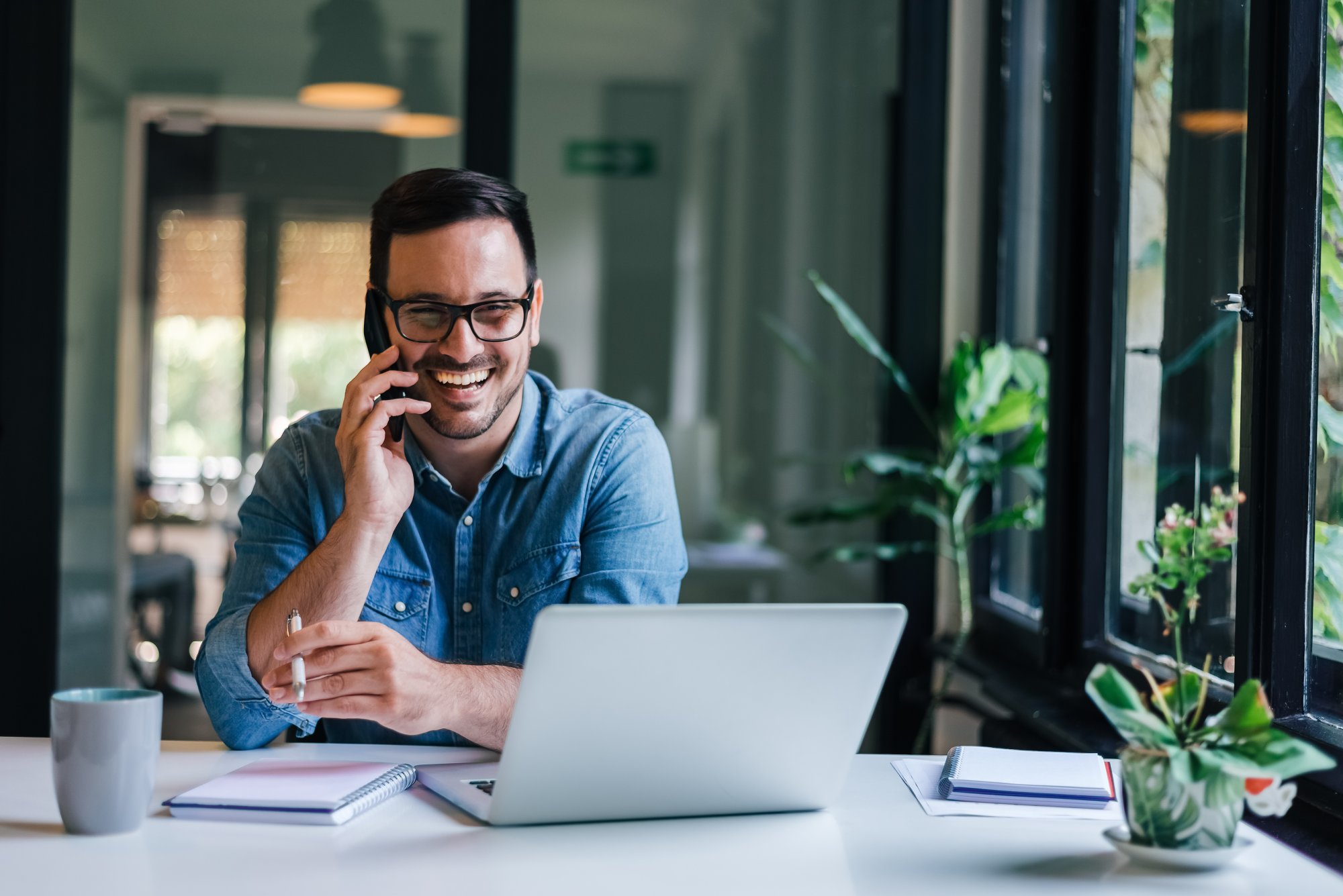 Entrepreneur on Phone Call with Laptop A professional man wearing glasses and a denim shirt sits at a modern desk, smiling while taking
