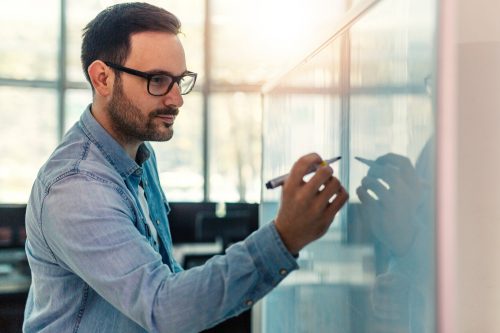 Professional Writing on Whiteboard by Window A man wearing glasses and a denim shirt writes on a glass whiteboard or transparent surface with