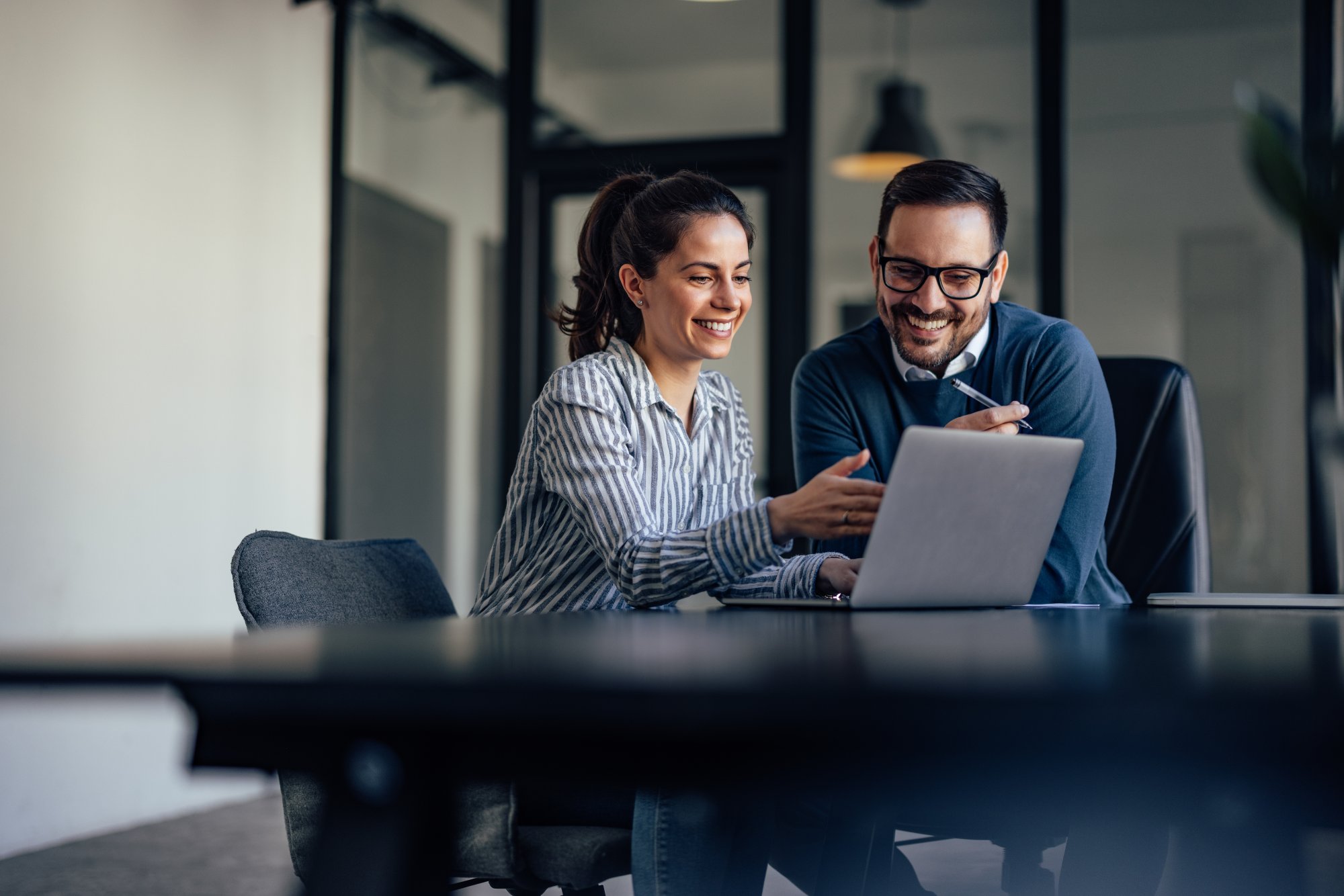 Two smiling professionals collaborate at a modern office desk, reviewing content on a laptop