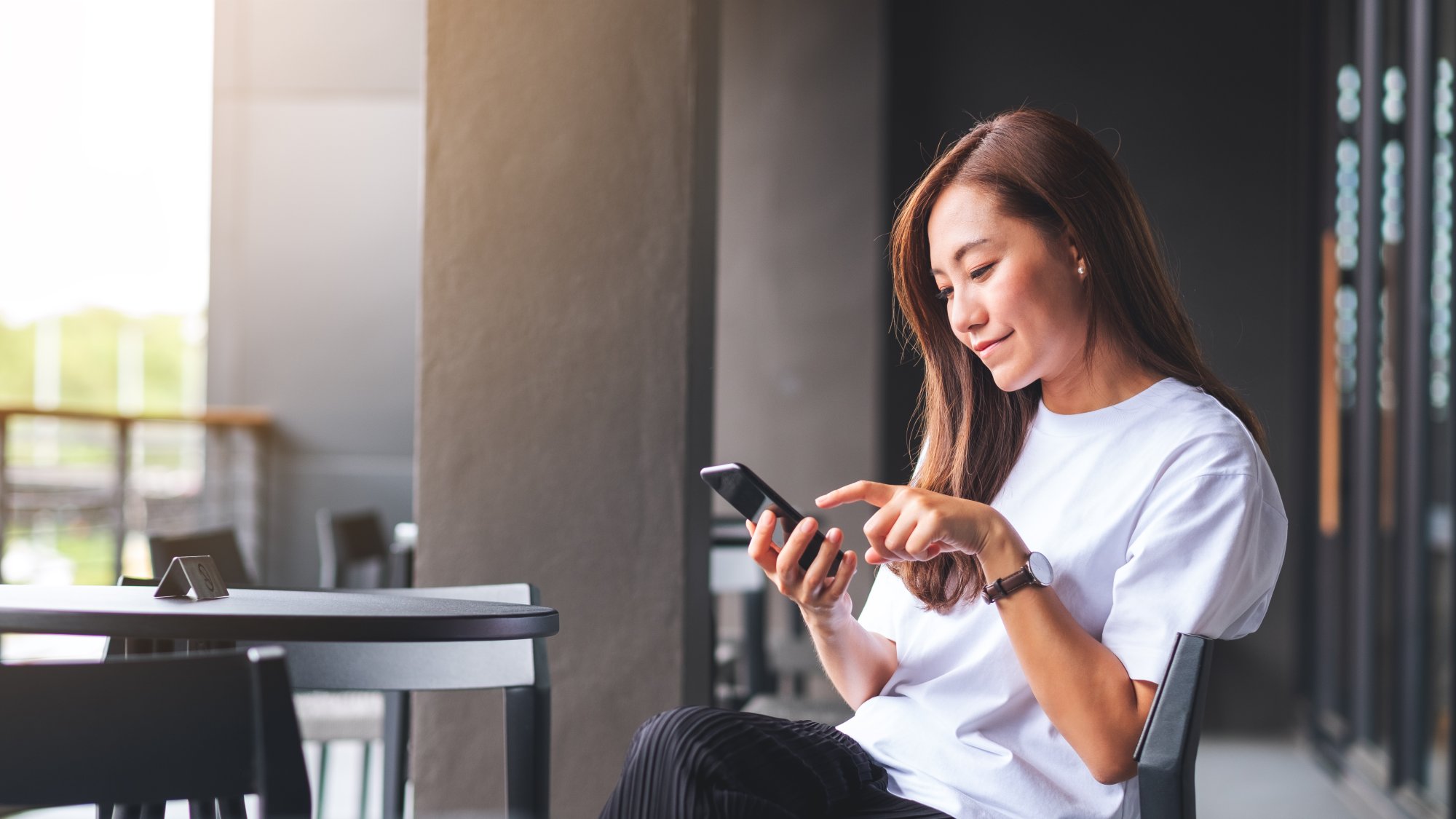 Woman Using Mobile Phone Outdoors A young Asian woman in a white t-shirt sits outdoors in a modern architectural setting, focused